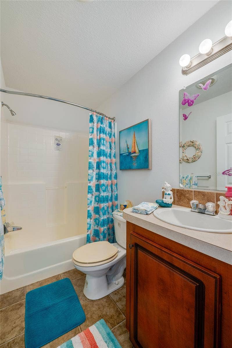 60 GOLDEN GATE CIRCLE, Port Orange, FL 32129 Full bathroom equipped with a wood vanity cabinet, mirror lighting fixture, toilet, and full tub-and-shower combination. Tile-patterned flooring adds texture to the space.