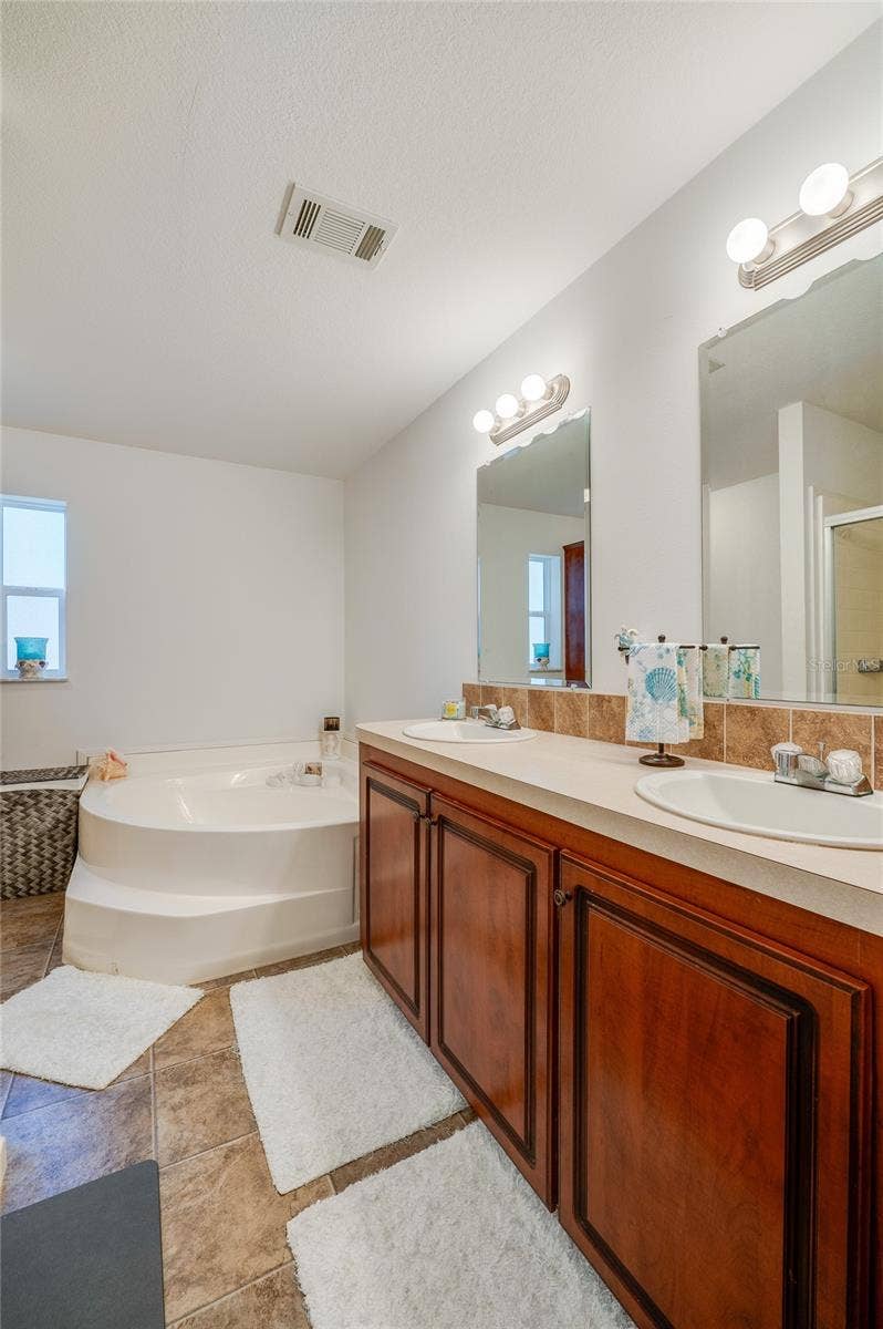 60 GOLDEN GATE CIRCLE, Port Orange, FL 32129 Ensuite primary bathroom featuring a double-sink vanity with wood cabinetry, bright vanity lighting, tile flooring, and a large corner soaking tub.