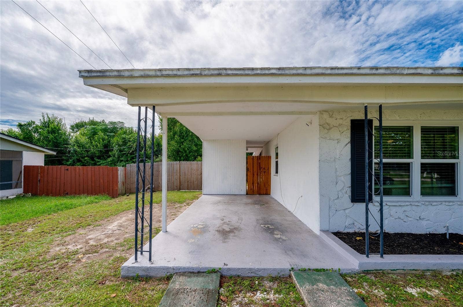 605 E NORMANDY BOULEVARD, Deltona, FL 32725 Covered concrete carport with wrought-iron support posts, a textured exterior wall finish, and a gated side pathway.