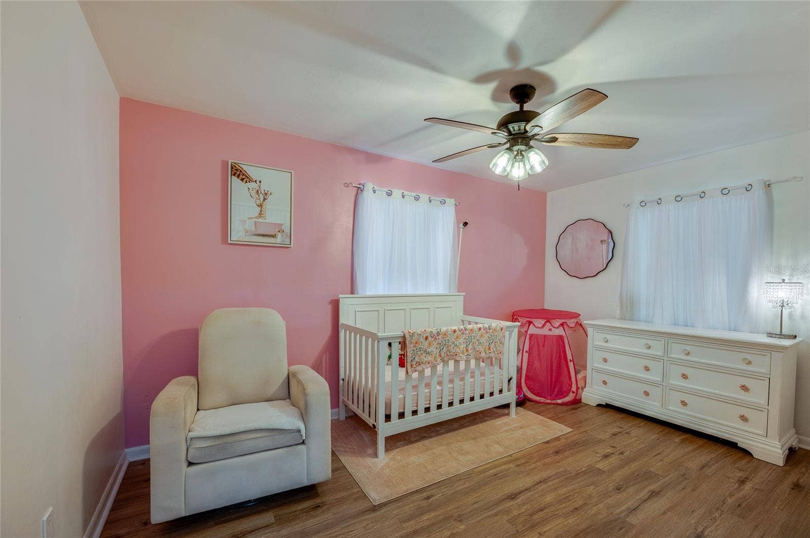 605 E NORMANDY BOULEVARD, Deltona, FL 32725 Bright bedroom area highlighted by warm LVP flooring, soft pink accent walls, a ceiling fan, and ample natural light from two windows.