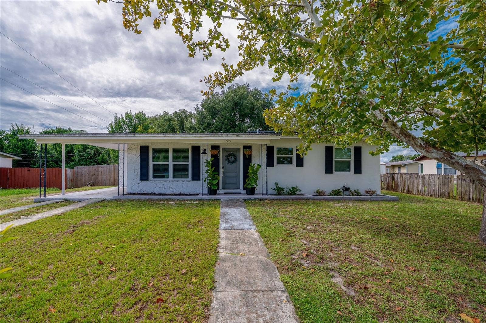605 E NORMANDY BOULEVARD, Deltona, FL 32725 Charming single-story home exterior featuring crisp white siding, dark accent shutters, a covered carport, and a paved walkway crossing a spacious front lawn.