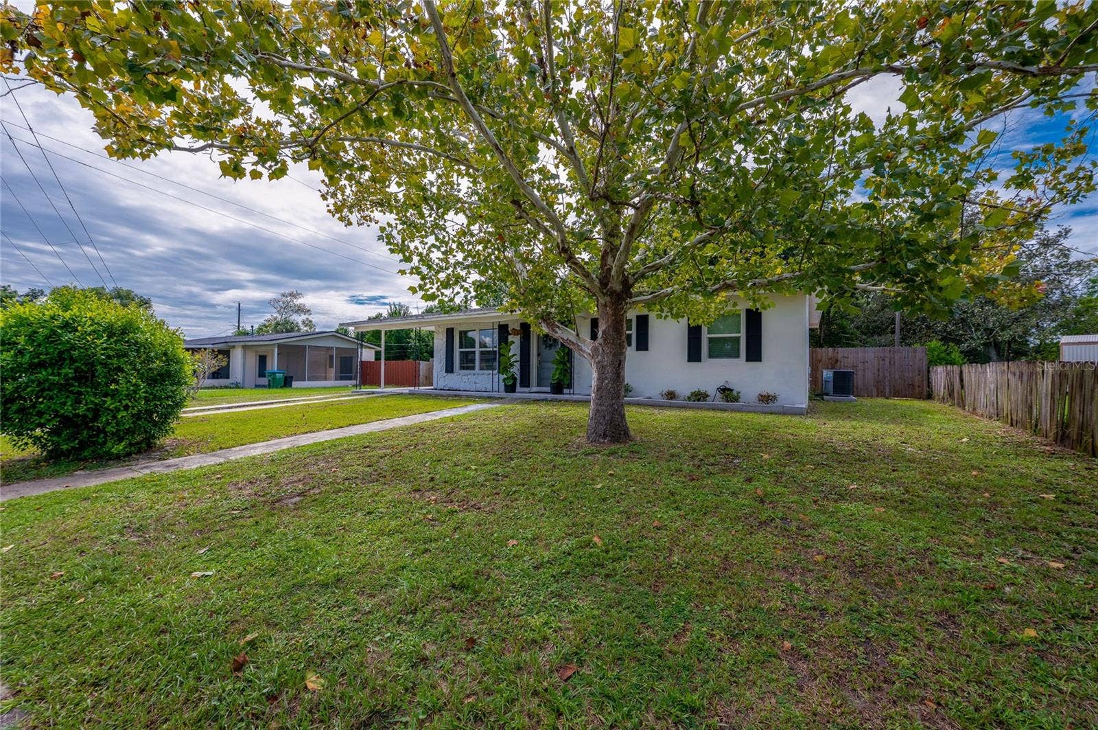 605 E NORMANDY BOULEVARD, Deltona, FL 32725 Lush front yard shaded by a large leafy tree, featuring a paved walkway leading to the home's single-story facade with dark window shutters.
