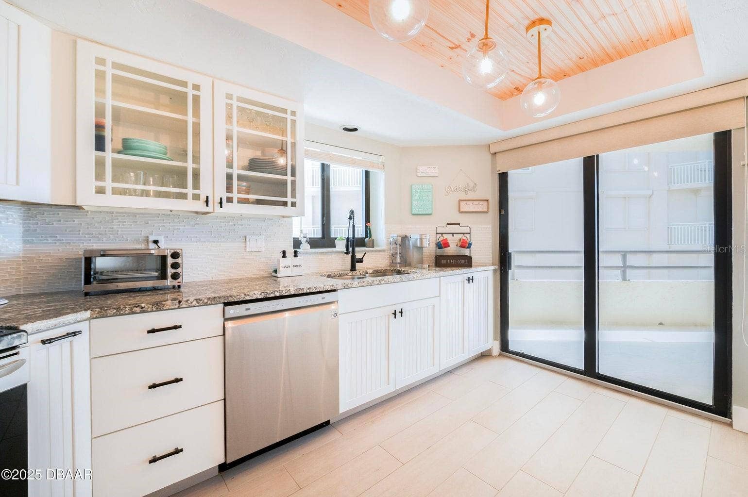 Beautifully appointed kitchen featuring white cabinetry, granite countertops, a stylish tile backsplash, and glass-front upper cabinets. A wood-accented tray ceiling with globe pendant lighting adds warmth and architectural character.