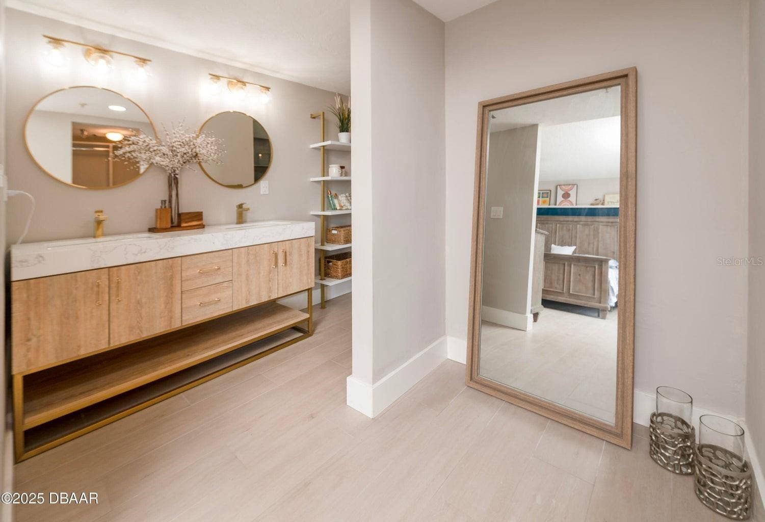 This beautifully updated primary bathroom features a double-sink vanity with wood-tone cabinetry, gold-tone fixtures, round mirrors, and contemporary lighting for a sophisticated coastal look.