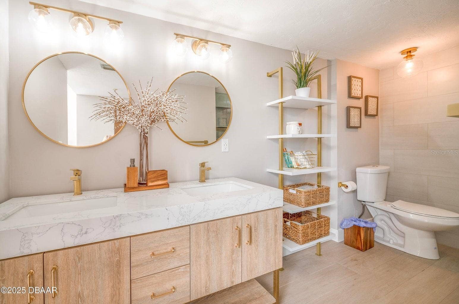 The spacious double vanity showcases marble-look countertops with integrated sinks, warm wood-tone cabinetry, stylish gold accents, and open shelving for convenient storage.