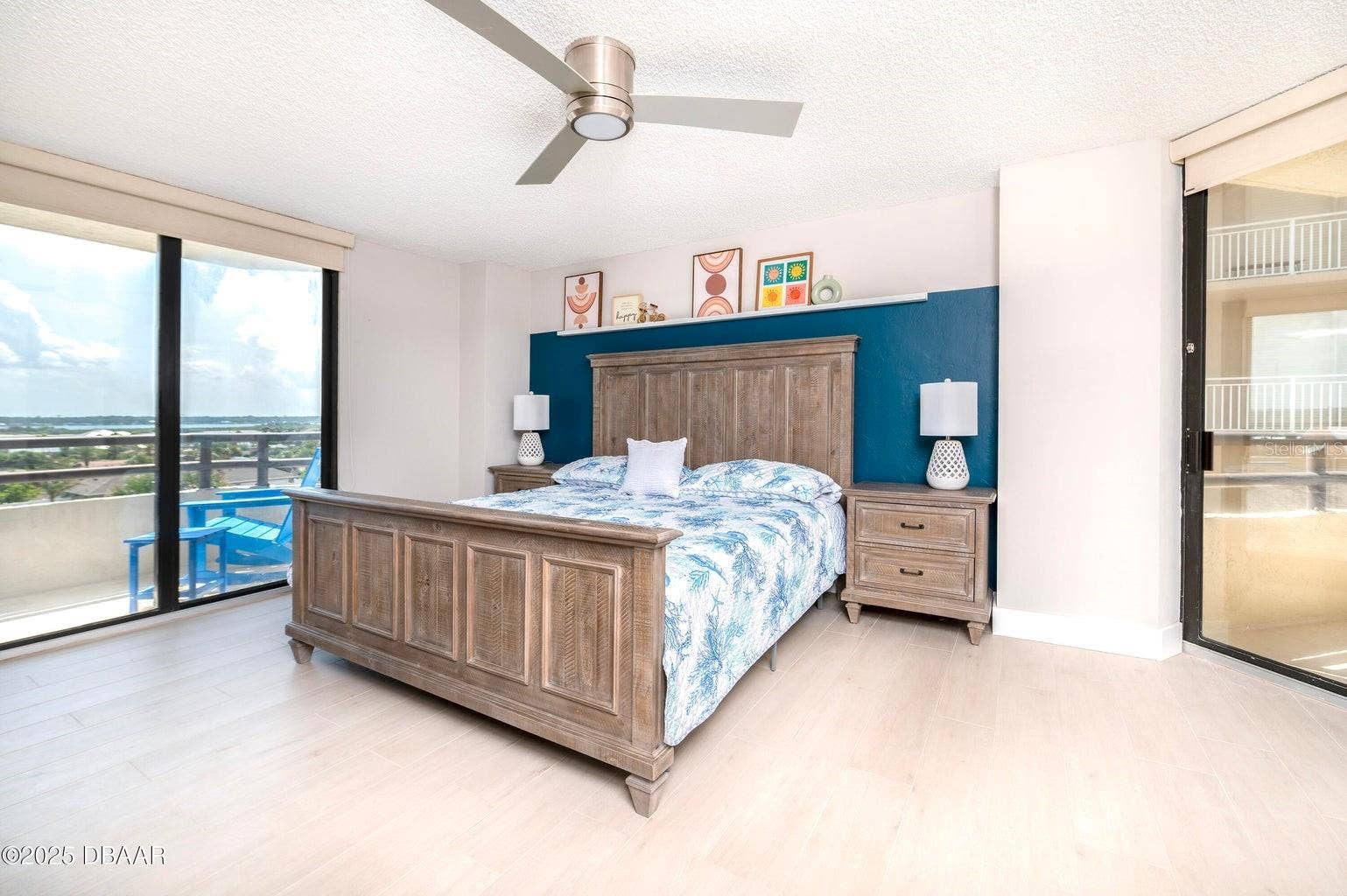 This spacious primary suite features a striking blue accent wall, contemporary ceiling fan, and sliding glass doors opening to a private balcony with scenic inland views.