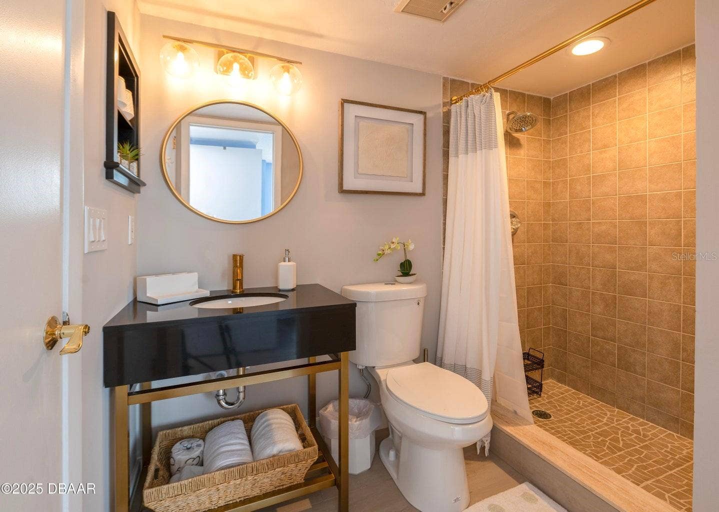 This stylish guest bathroom features a modern black vanity with gold-tone accents, a round mirror, updated lighting, and a tiled walk-in shower with a rainfall showerhead.