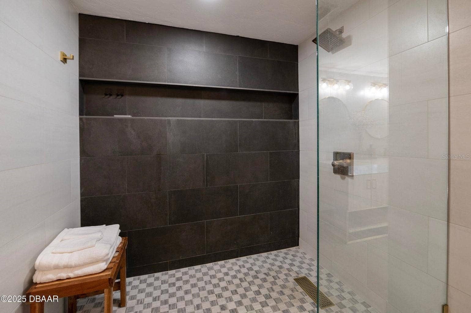 This spacious walk-in shower features a striking dark tile accent wall, contrasting light tile surrounds, a rainfall showerhead, built-in storage niche, and sleek glass partition for a contemporary, spa-inspired feel.