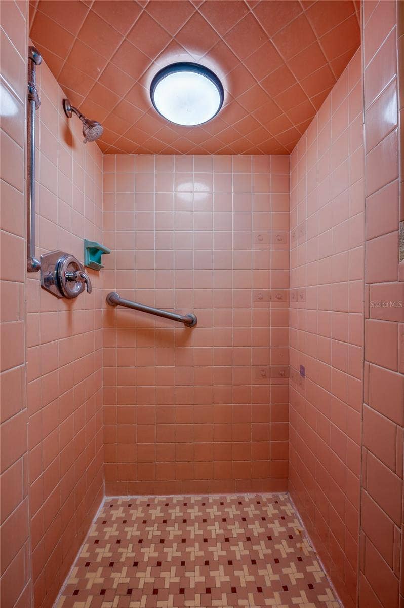 Dedicated walk-in shower stall accented with blush-pink wall tiles, a arched tile ceiling, and matching patterned mosaic floor tile. The space includes a handheld showerhead attachment and integrated wall grab bar.