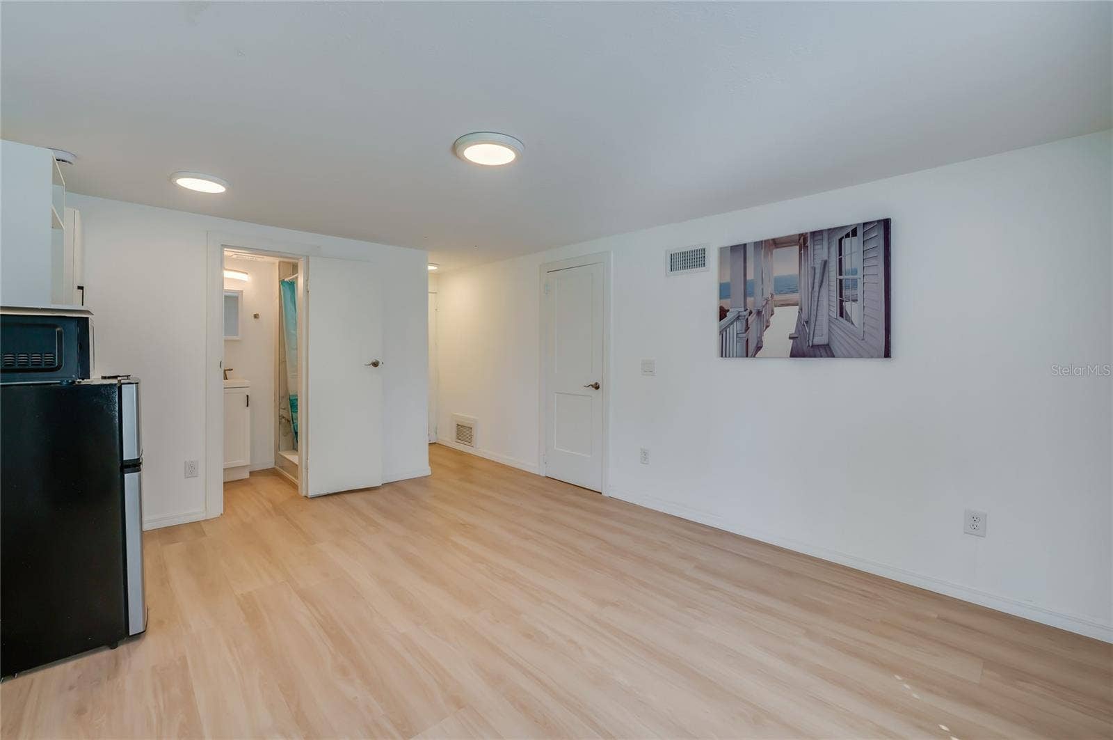 Bright indoor living space showcasing crisp white walls, LVP flooring, and recessed ceiling lights. An open doorway leads directly toward the bathroom area.