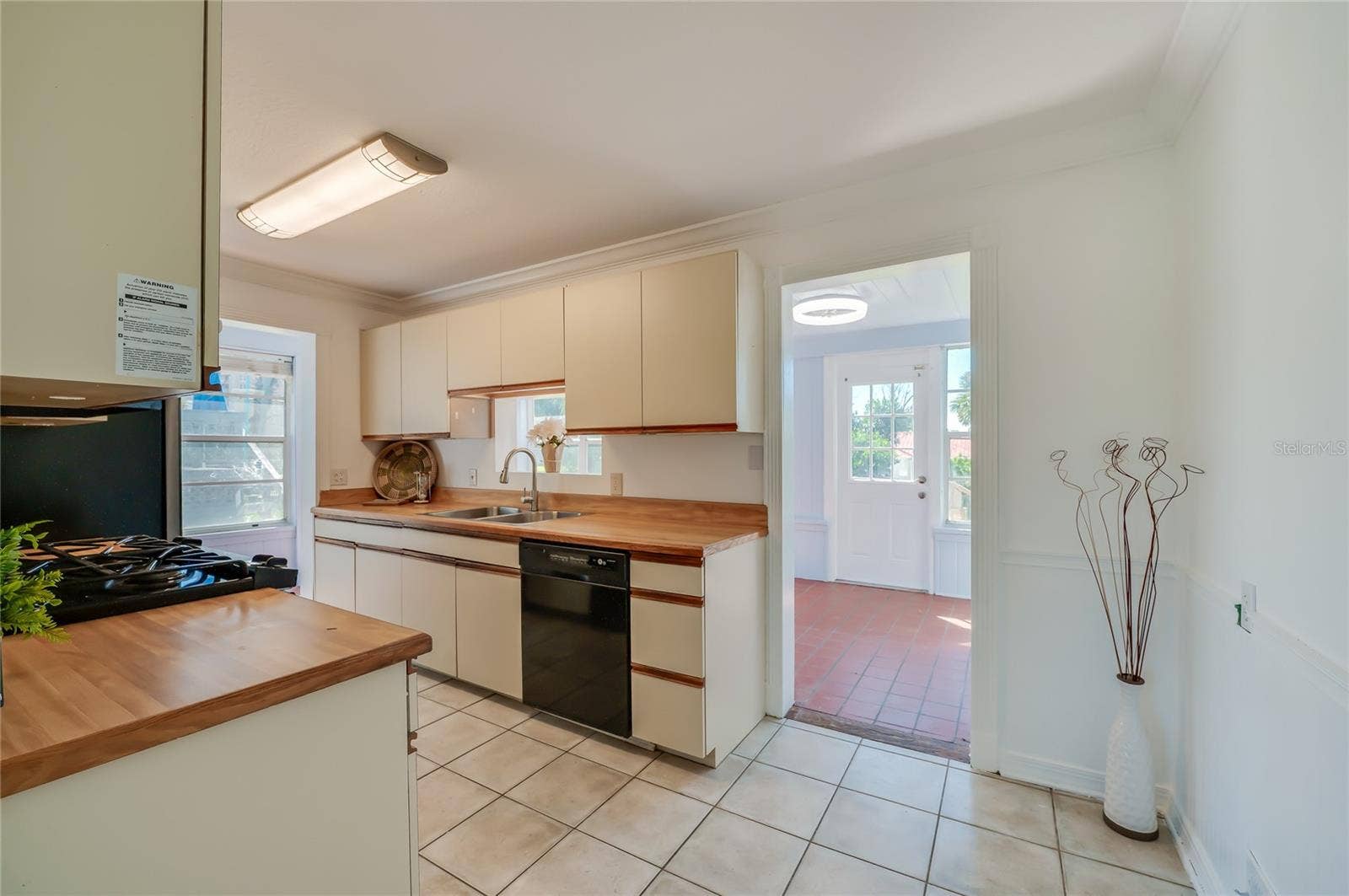Functional kitchen featuring butcher block-style countertops, classic cabinetry, tile flooring, and integrated black appliances. An adjacent open doorway provides direct access to a sunlit room with terracotta tile floors.