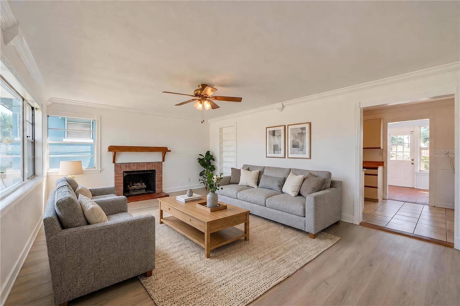 Virtually Staged - Spacious living area showcasing light LVP flooring, crown molding, and a charming brick fireplace with a wooden mantel. Large windows allow plenty of natural light, and an open doorway leads toward the kitchen and sunroom areas.