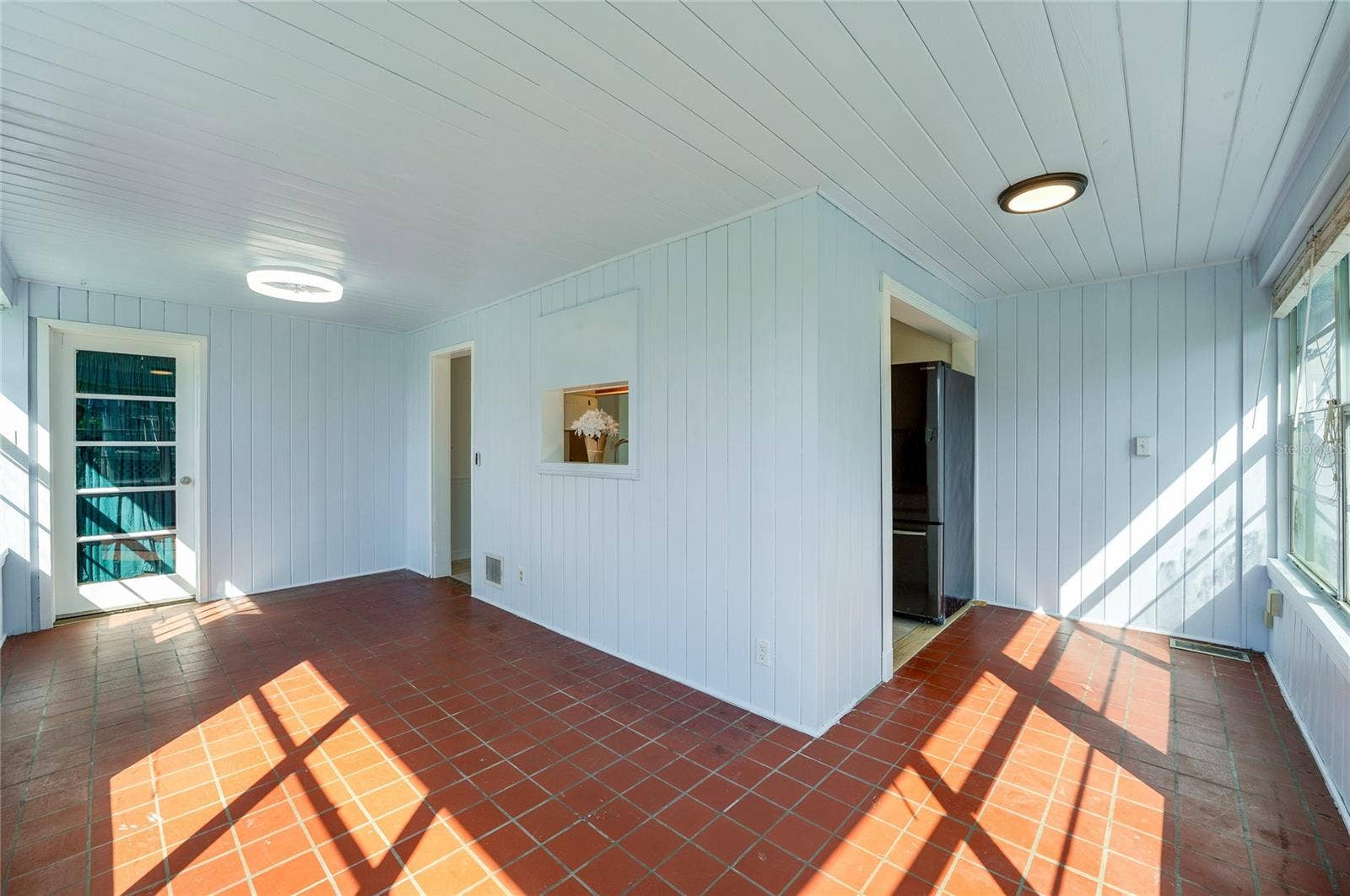 Sun-filled dining area and breakfast nook showcasing red tile flooring, modern flush-mount lighting, and white paneled walls. Large windows fill the space with natural light while direct door access leads outdoors.