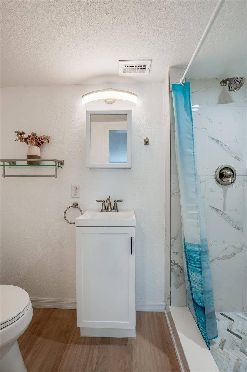 Compact bathroom area highlighting a sleek white vanity, a medicine cabinet, and bright wall sconce lighting. A walk-in shower sits alongside LVP flooring.