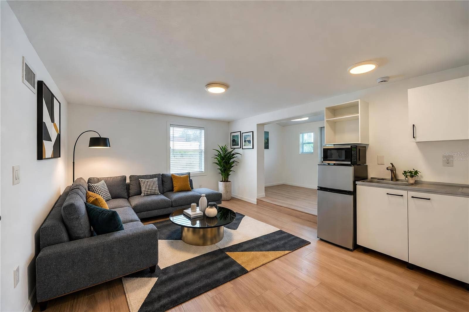 Virtually Staged - Open studio in law suite boasting modern kitchenette amenities including a compact refrigerator, microwave, sink, and low-profile cabinetry. Light LVP flooring and recessed overhead lights create a clean, contemporary feel.