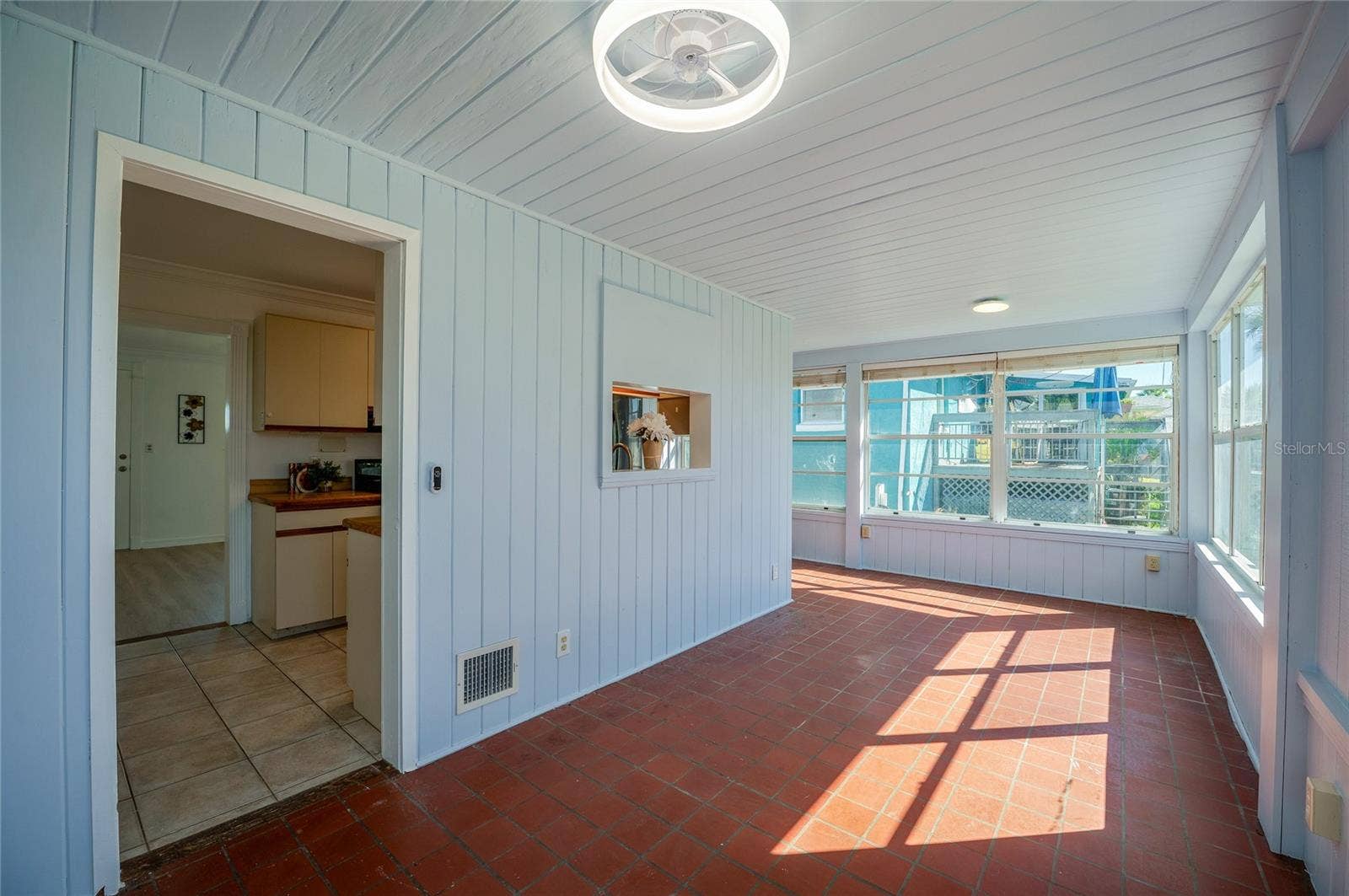 Dinning area featuring light blue wood-paneled walls, warm terracotta floor tiles, and abundant wrap-around windows. A passthrough window connects back to the kitchen for added convenience.