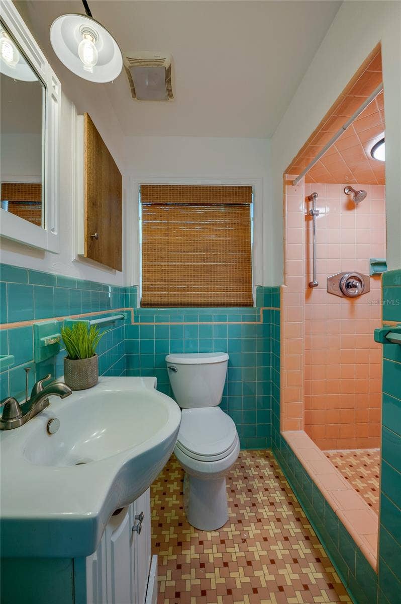 Vibrant vintage-style bathroom featuring turquoise wall tile, patterned geometric floor tile, and a matching pink-tiled walk-in shower stall. A white pedestal-style vanity with modern fixtures adds functional contrast.