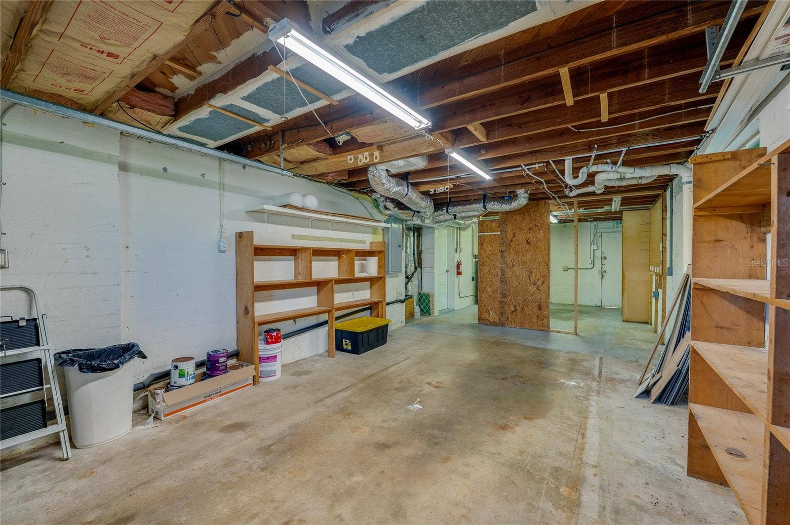 Expansive lower-level workshop or storage area featuring concrete flooring, exposed ceiling joists with overhead strip lighting, and built-in wooden shelving units.