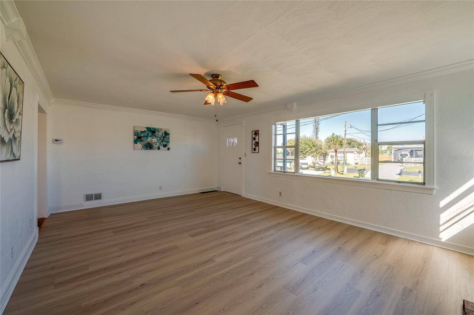 Bright living room highlighting smooth neutral walls, a ceiling fan with light fixtures, and a expansive front window overlooking the neighborhood. A side doorway connects seamlessly to the rest of the home's interior layout.