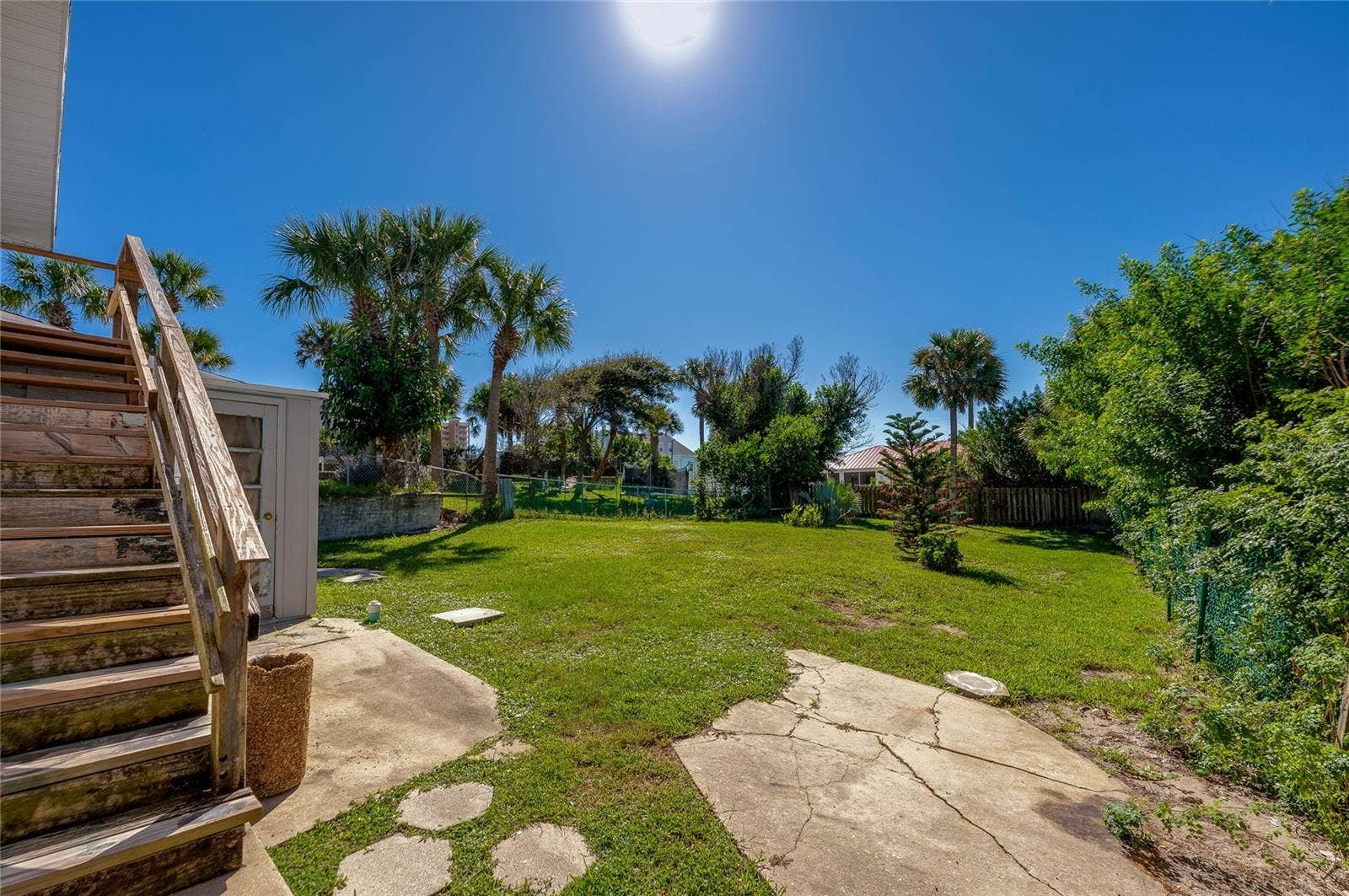 Spacious outdoor backyard featuring a grassy lawn, concrete patio stepping stones, and exterior wooden stairs leading up the residence. Lush tropical foliage and palm trees border the sunny yard.
