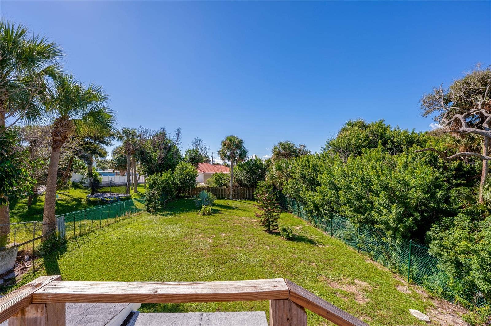 Spacious grassy backyard surrounded by lush palm trees, mature greenery, and secure fencing. An elevated wooden deck frame offers views across the entire lawn.