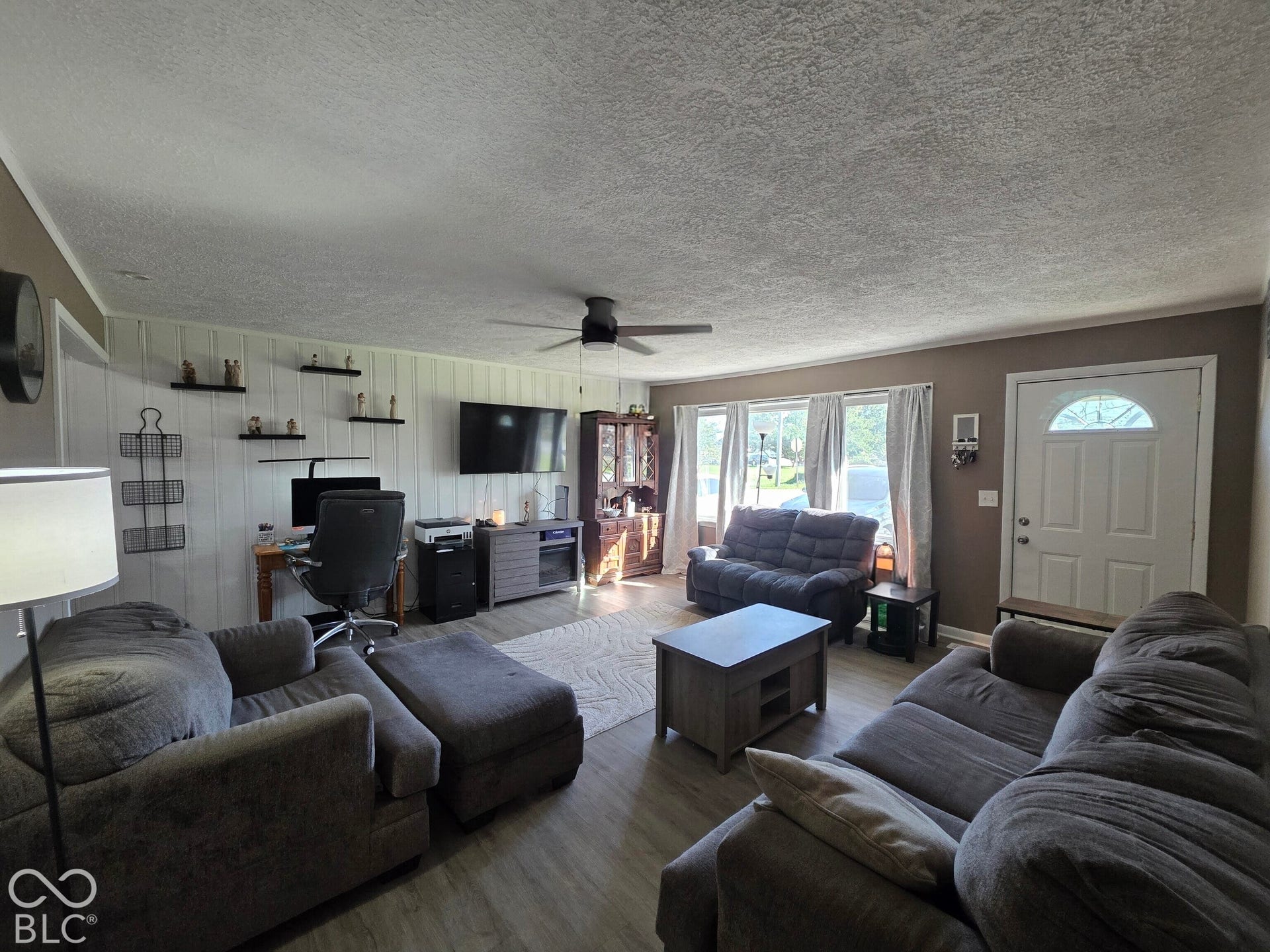 Living area featuring vertical board-and-batten accent wall, wood-finish flooring, ceiling fan, and large windows with natural lighting