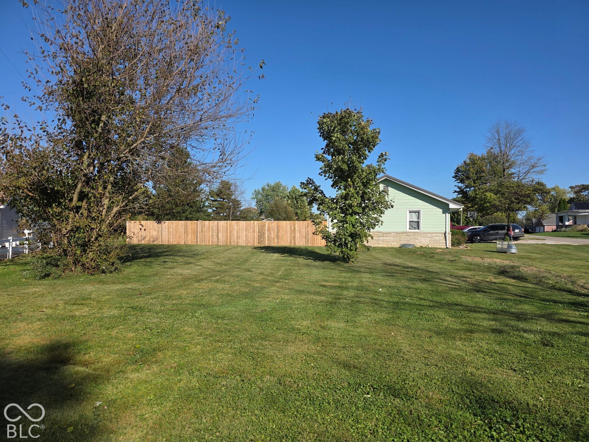 Expansive grassy lot featuring mature trees, wood privacy fencing, and exterior stone foundation detailing