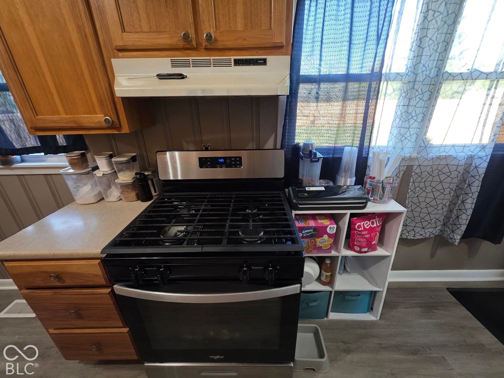 Stainless steel gas range featuring continuous cast-iron grates and an integrated range hood, set within a kitchen area with wood-finish cabinetry and neutral-toned countertops