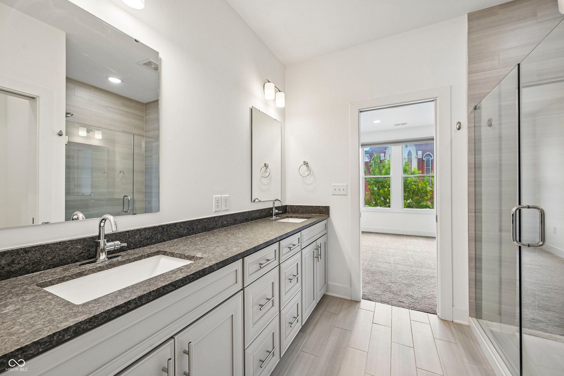 Bathroom featuring a dual vanity with a dark countertop and undermount sinks, a glass-enclosed shower with tile surround, and wood-finish flooring