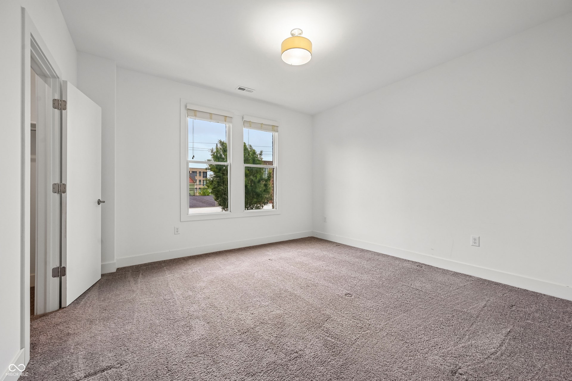 Spacious interior room featuring plush gray carpeting, white walls, two large windows, a ceiling-mounted light fixture, and a white door with dark hardware