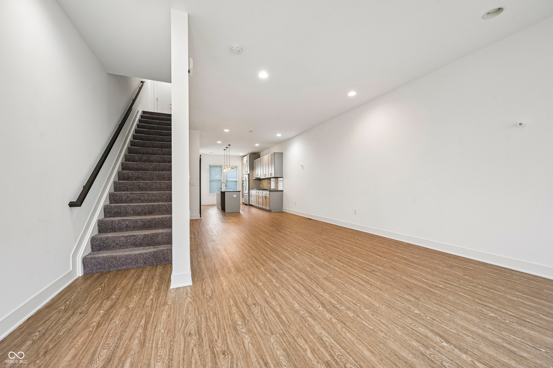 Open-concept living space featuring wood-finish flooring, recessed lighting, white walls, a carpeted staircase with a black handrail, and a kitchen with light-colored cabinetry and a central island