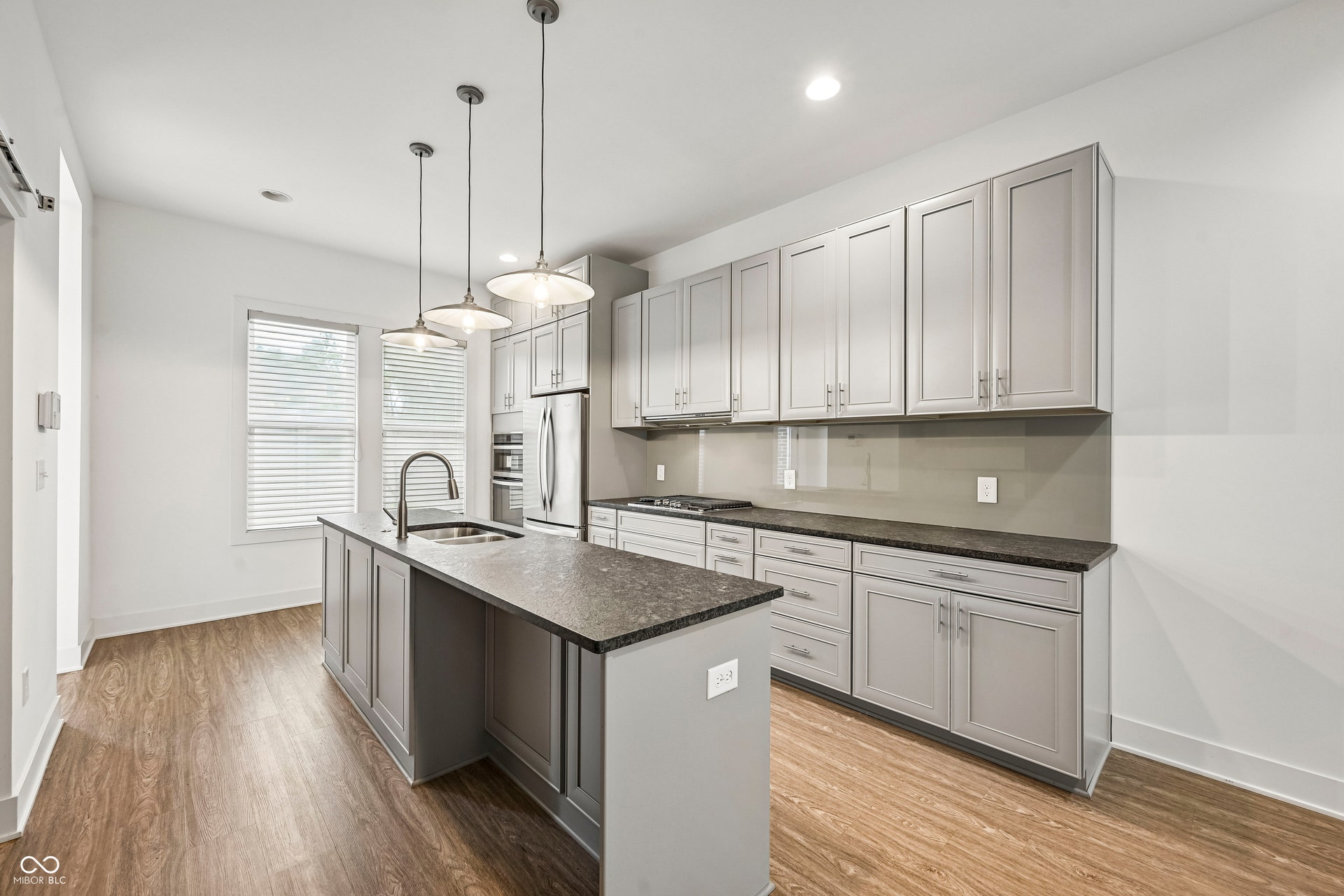 Spacious kitchen featuring an island with an undermount sink, pendant lighting, stainless steel appliances, light gray cabinetry, and stone countertops