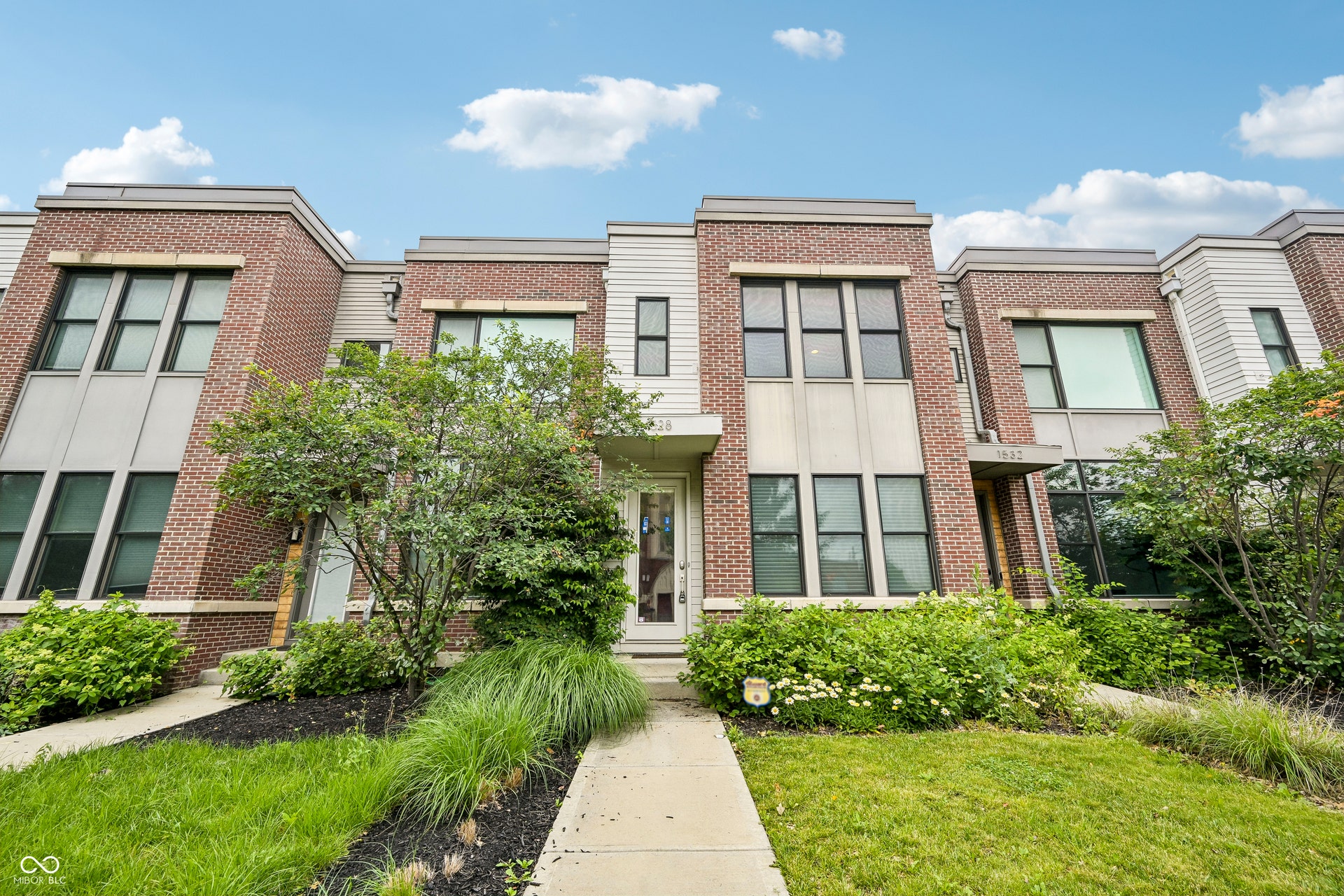 Contemporary townhome exterior featuring a brick and siding facade, multiple large windows, a paved walkway, and maintained landscaping with mature shrubs and ornamental grasses