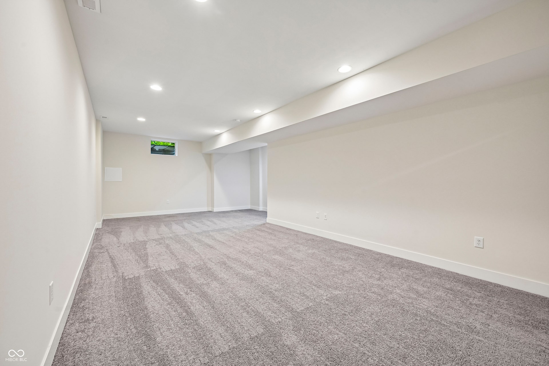Spacious room featuring neutral carpeting, recessed lighting, light-toned walls, white baseboards, and a window providing natural light