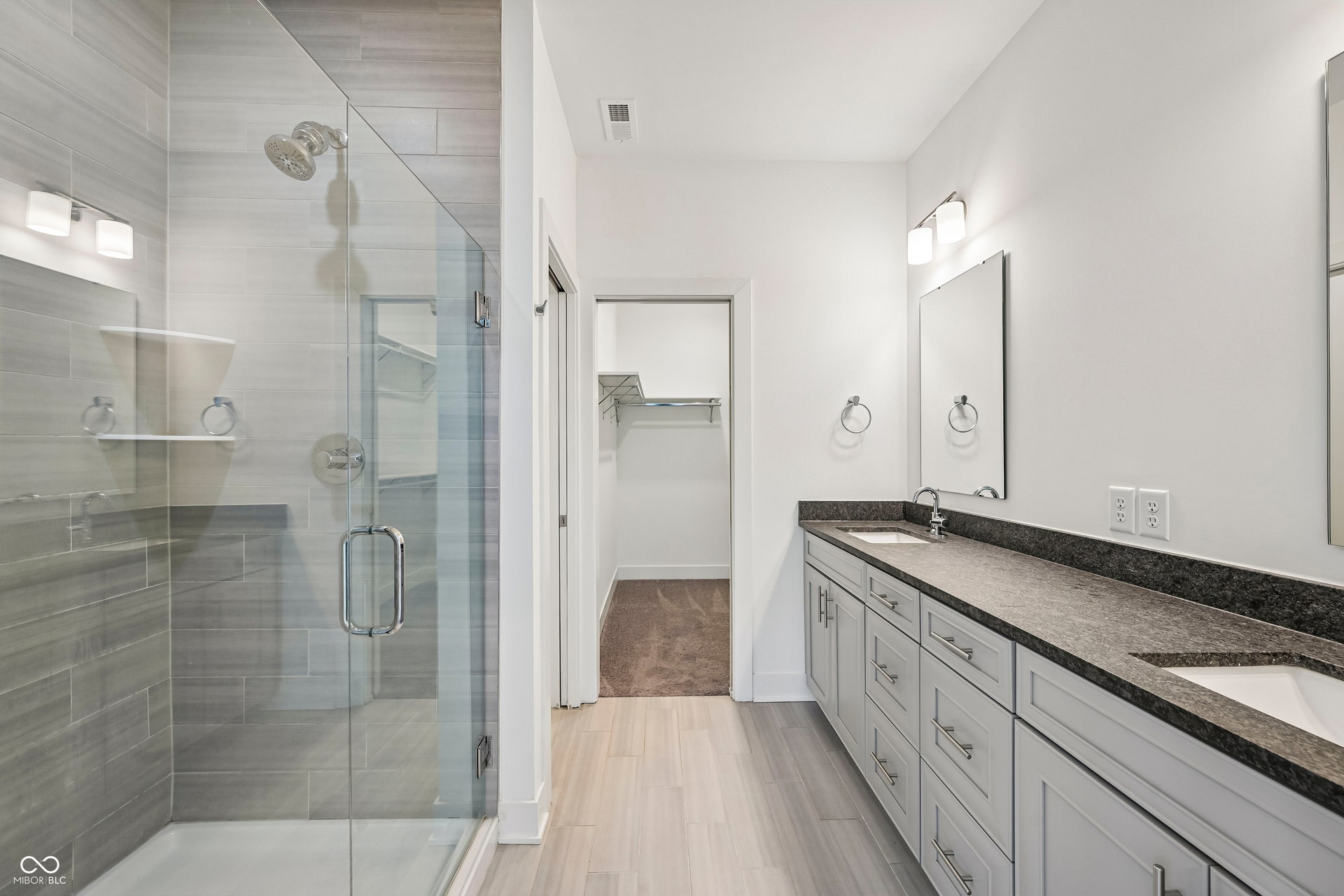 Spacious bathroom featuring a glass-enclosed shower with dual shower heads, a dual vanity with undermount sinks and dark countertops, light wood-finish flooring, and a walk-in closet with built-in shelving