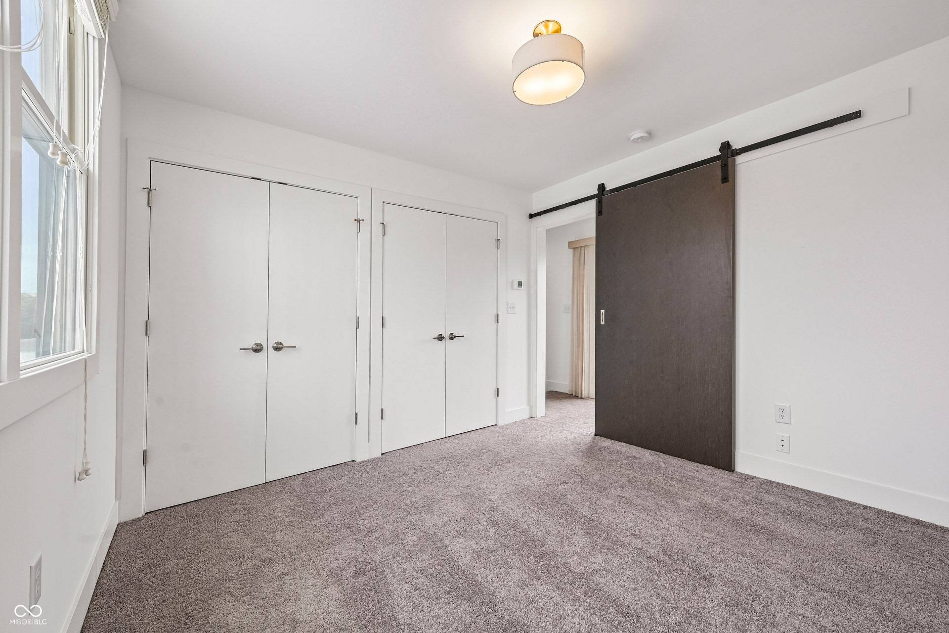 Spacious room featuring a dark barn door, light-toned carpeting, two white closet doors, a window with blinds, and a contemporary flush mount light fixture
