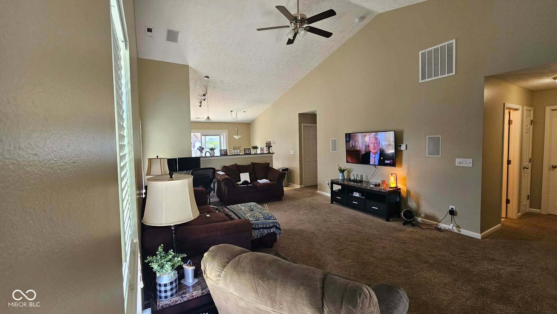 Expansive living room featuring vaulted ceilings, ceiling fan, carpet flooring, and a media wall with built-in speakers