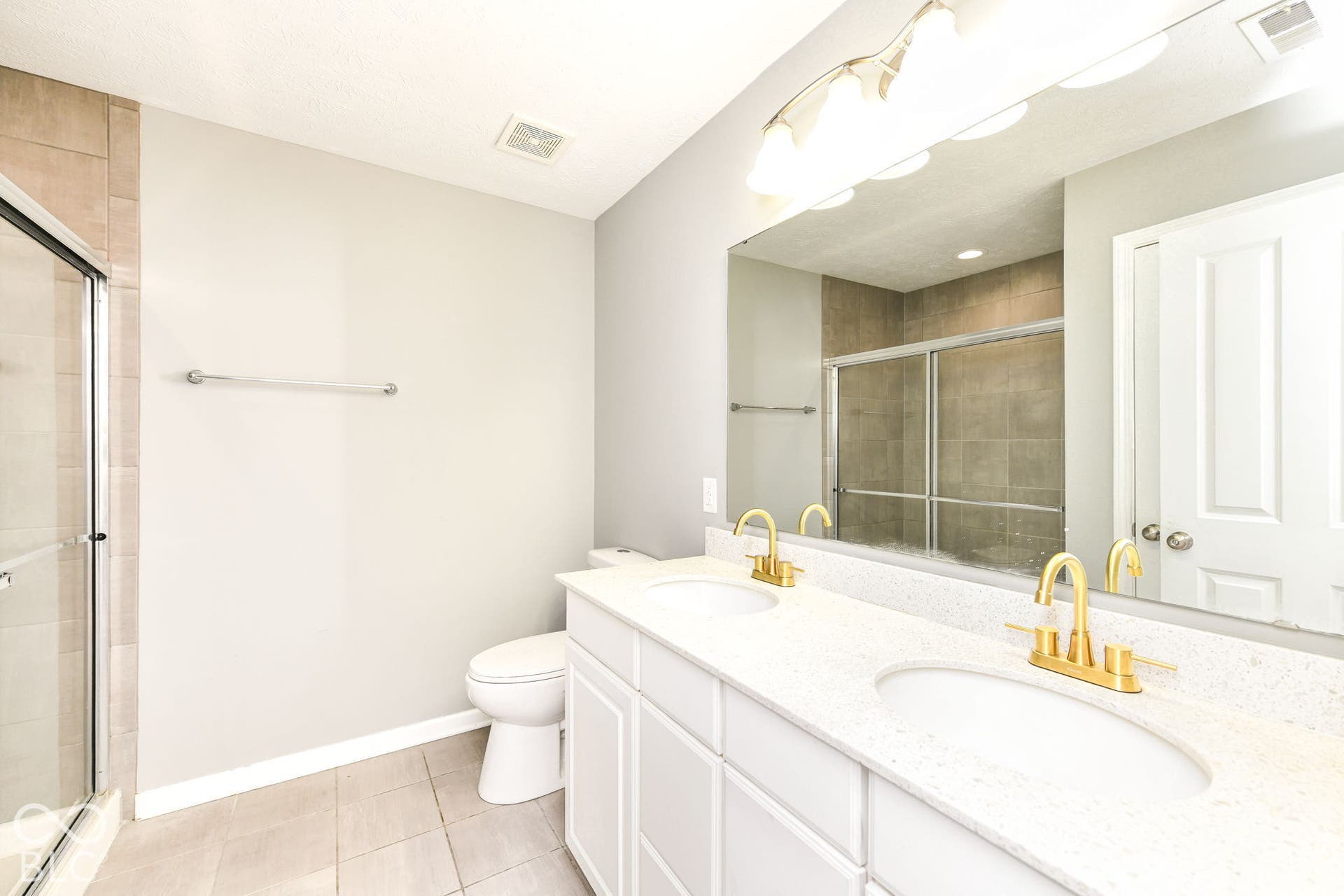 Dual vanity with integrated sinks, gold-finish fixtures, a large mirror, and an adjacent enclosed shower with tiled walls