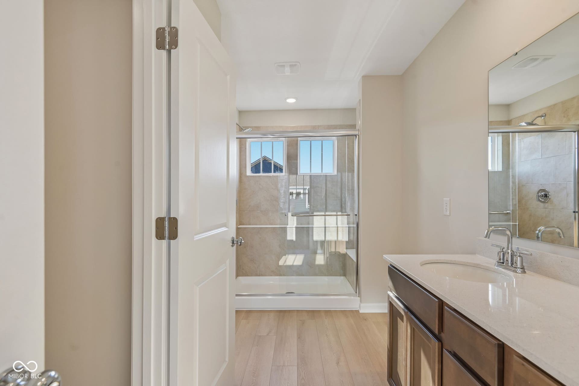 Bathroom featuring a frameless glass shower enclosure with tiled walls, a single vanity with a white countertop and undermount sink, and wood-finish flooring