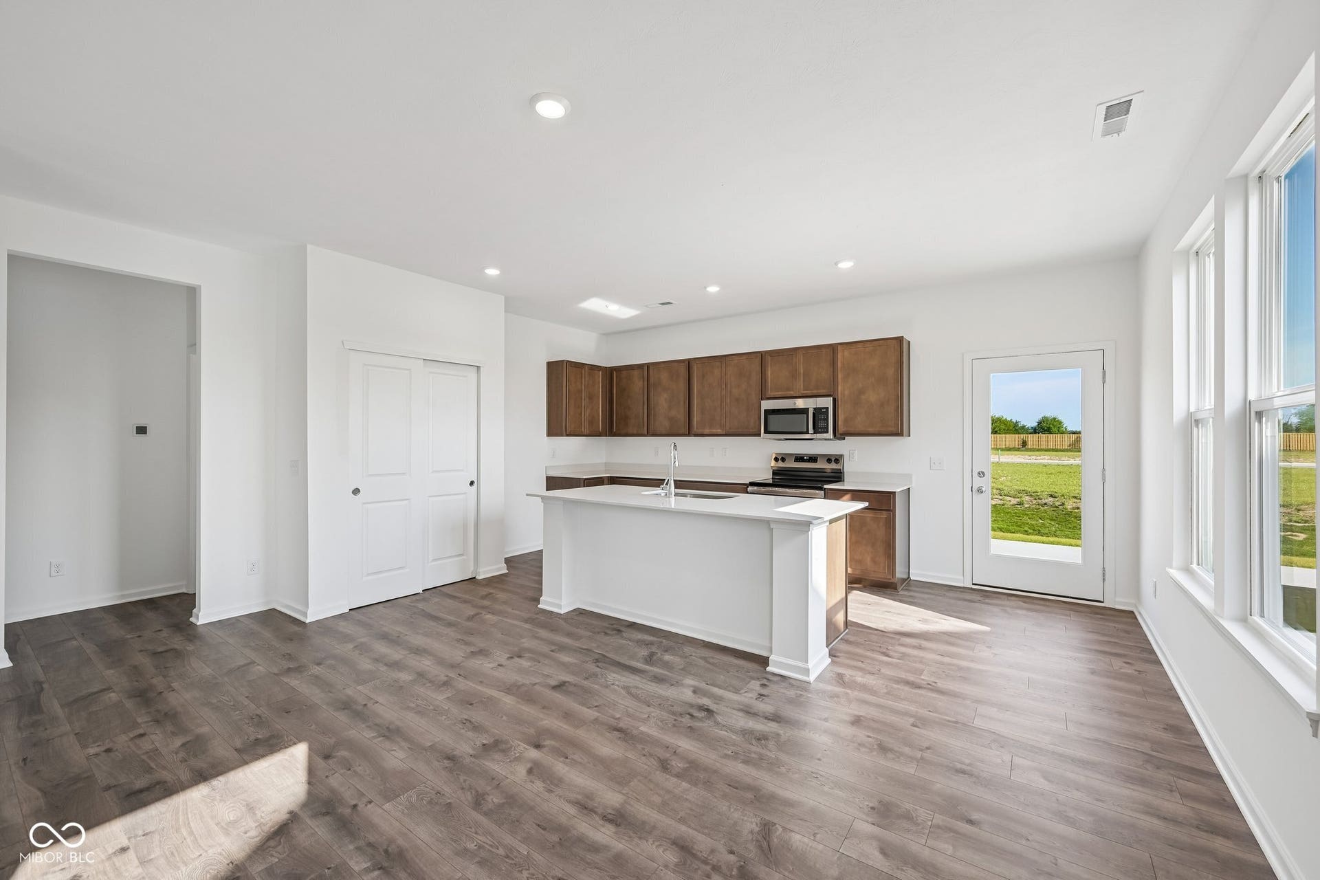 Open-concept kitchen with wood-finish flooring, white island, wood cabinetry, stainless steel appliances, and a full-light door