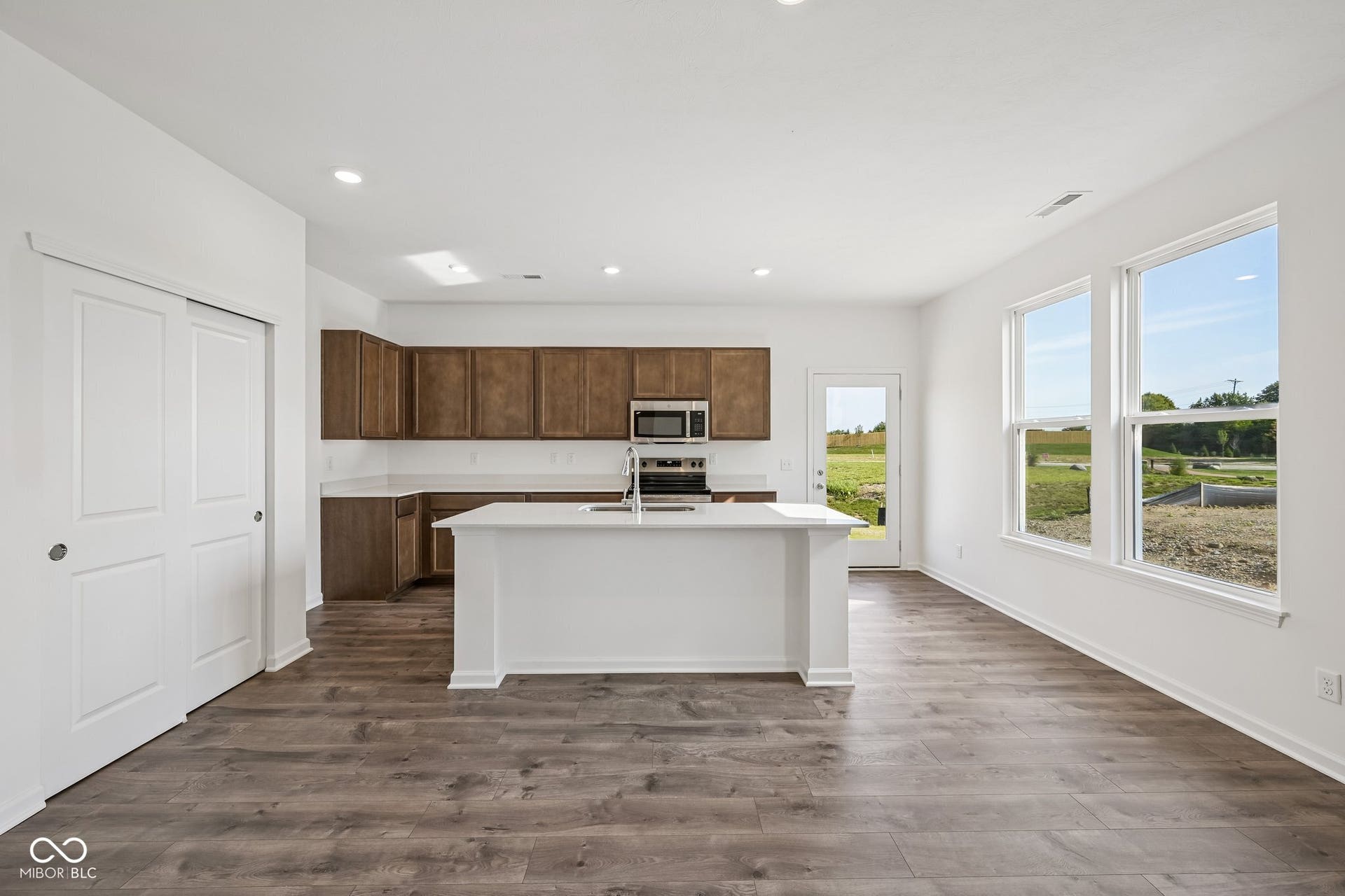 Open-concept living space with wood-finish flooring, a kitchen island with integrated sink, stainless steel appliances, and abundant natural light from multiple windows