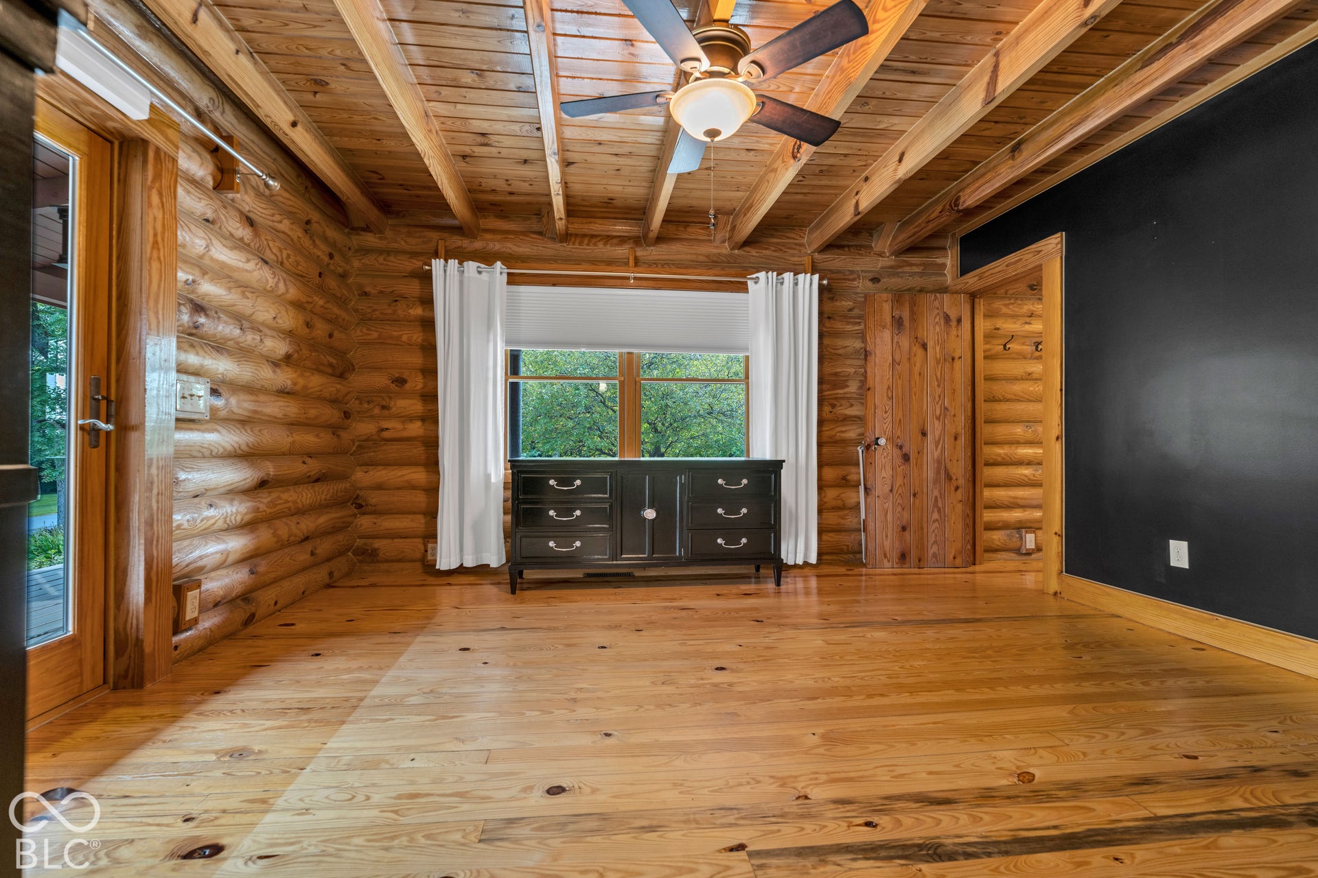 Exposed wood beam ceiling, wood-finish flooring, log wall construction, wood-frame window, and a dark accent wall