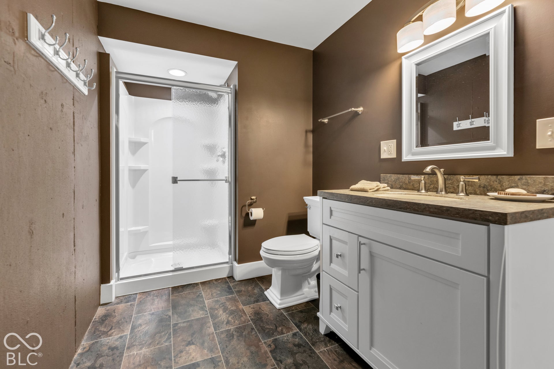 White vanity with a neutral-toned countertop and brushed nickel fixtures, glass-enclosed shower, tiled flooring, and a framed mirror with overhead lighting