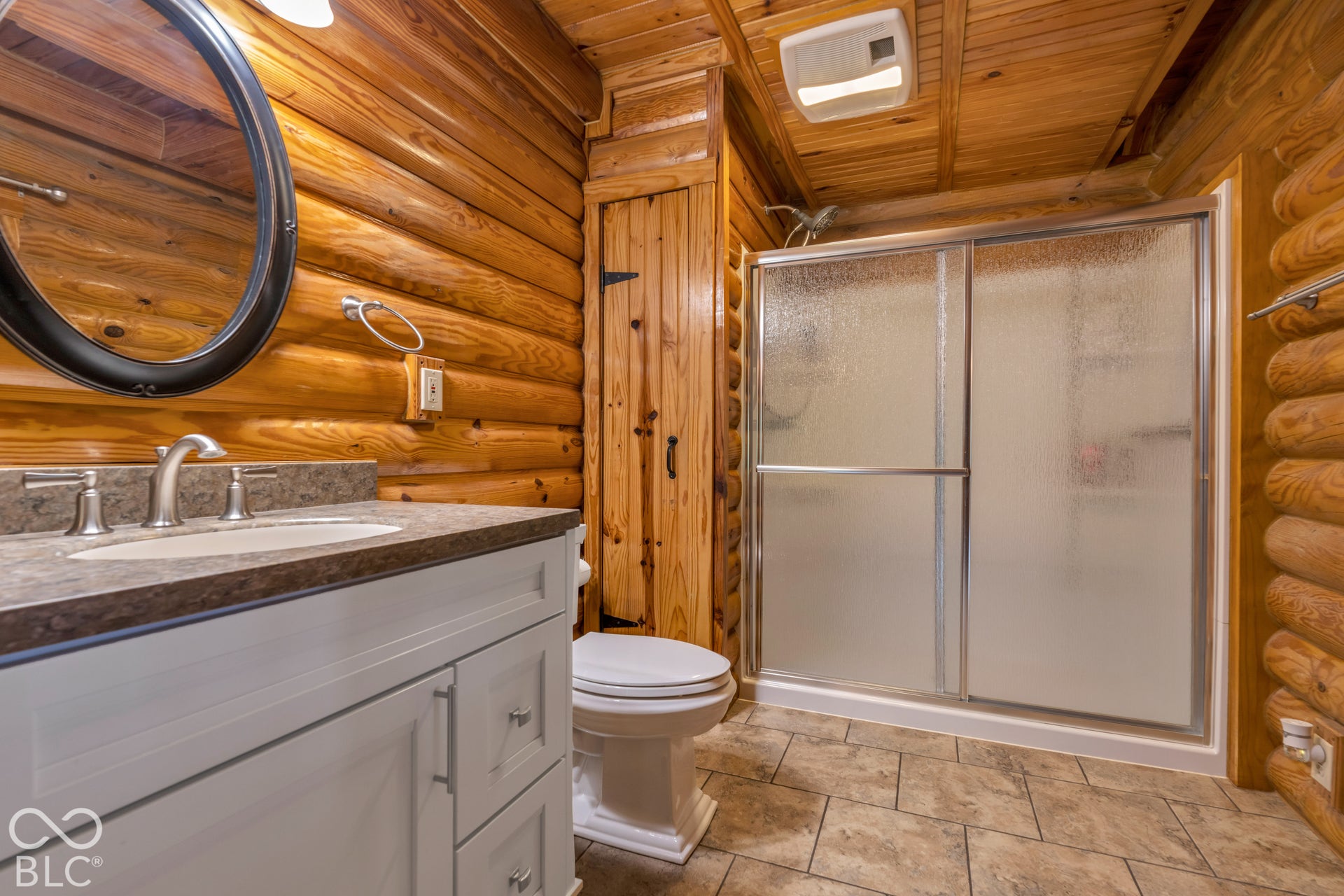 Log wall paneling, white vanity with stone-finish countertop, brushed nickel fixtures, and a shower with frosted glass doors