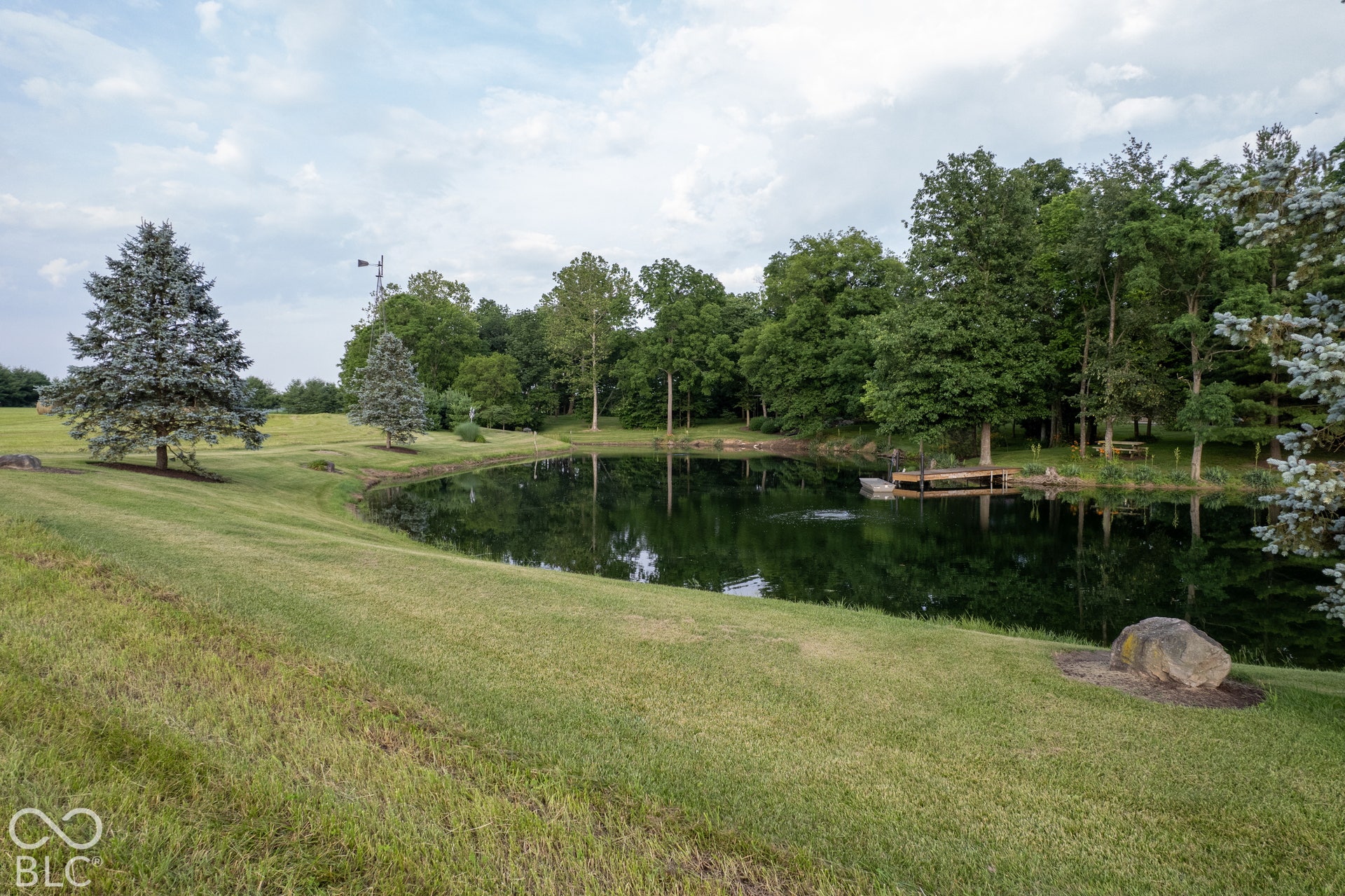 Pond featuring a wooden dock, surrounded by mature trees and manicured lawns