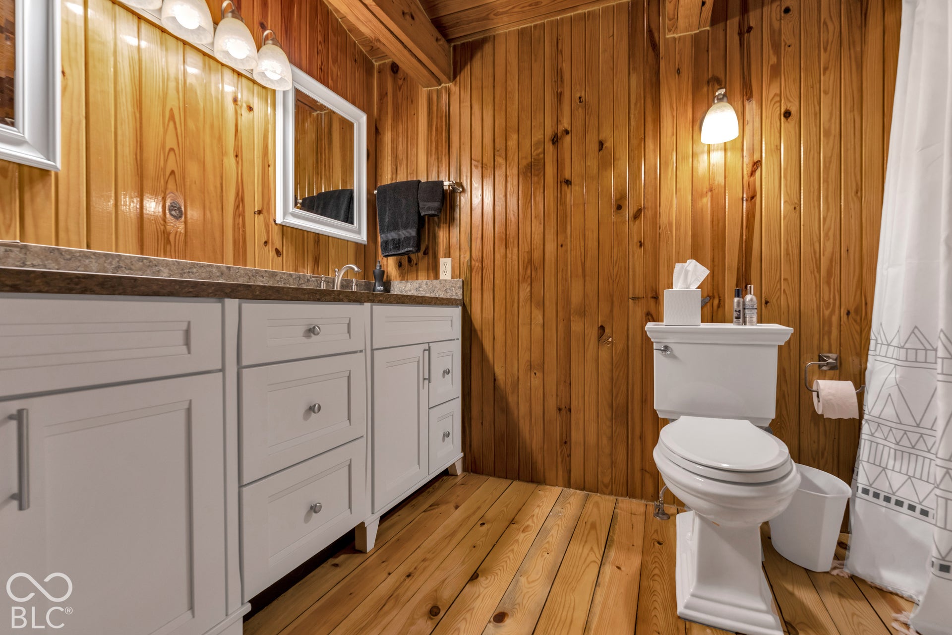 Wood-paneled walls and ceiling, wood-finish flooring, dual vanity with light cabinetry, and a countertop with integrated sinks