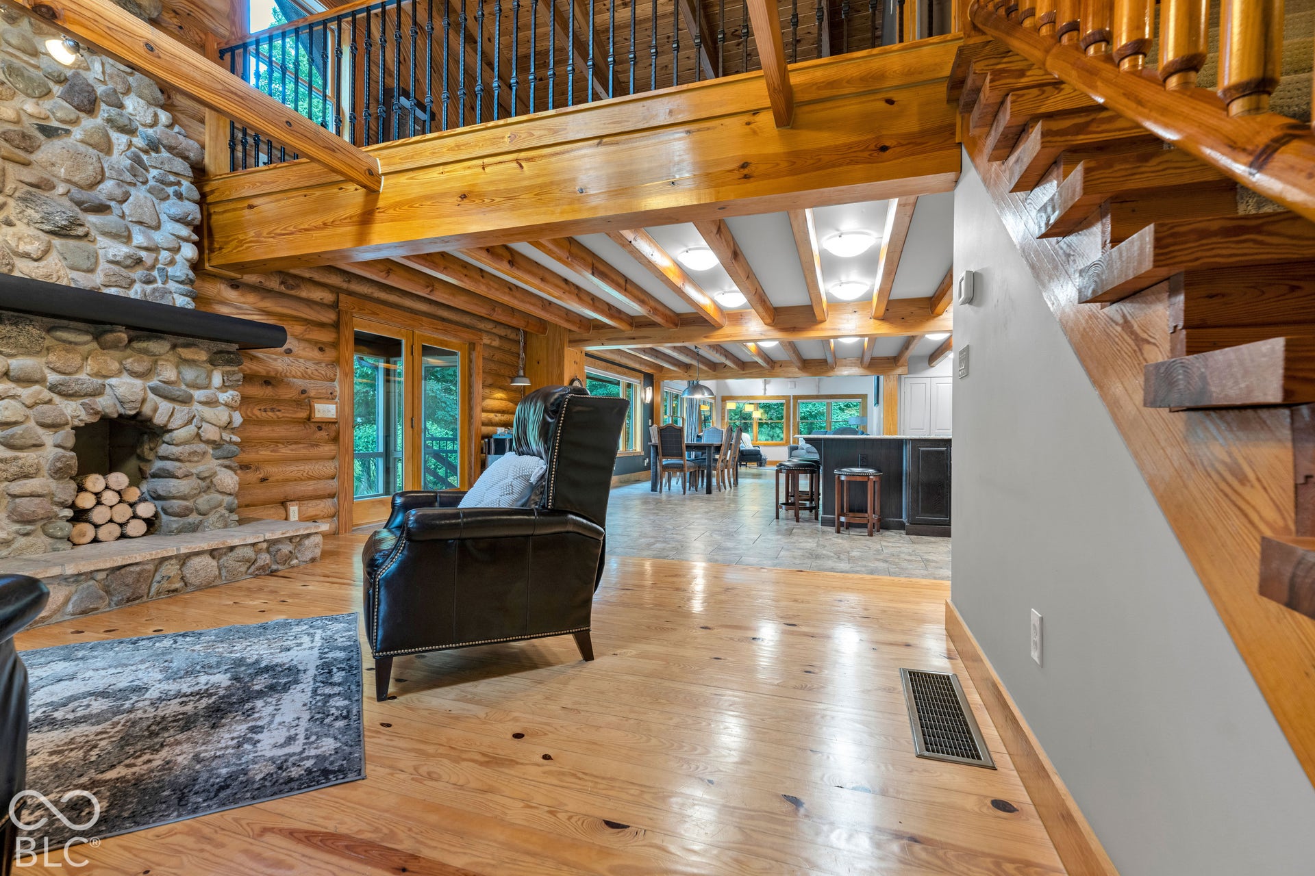 Stone-faced fireplace with a substantial mantel, log wall construction, exposed timber ceiling beams, wood-finish flooring, and a wooden staircase with balusters