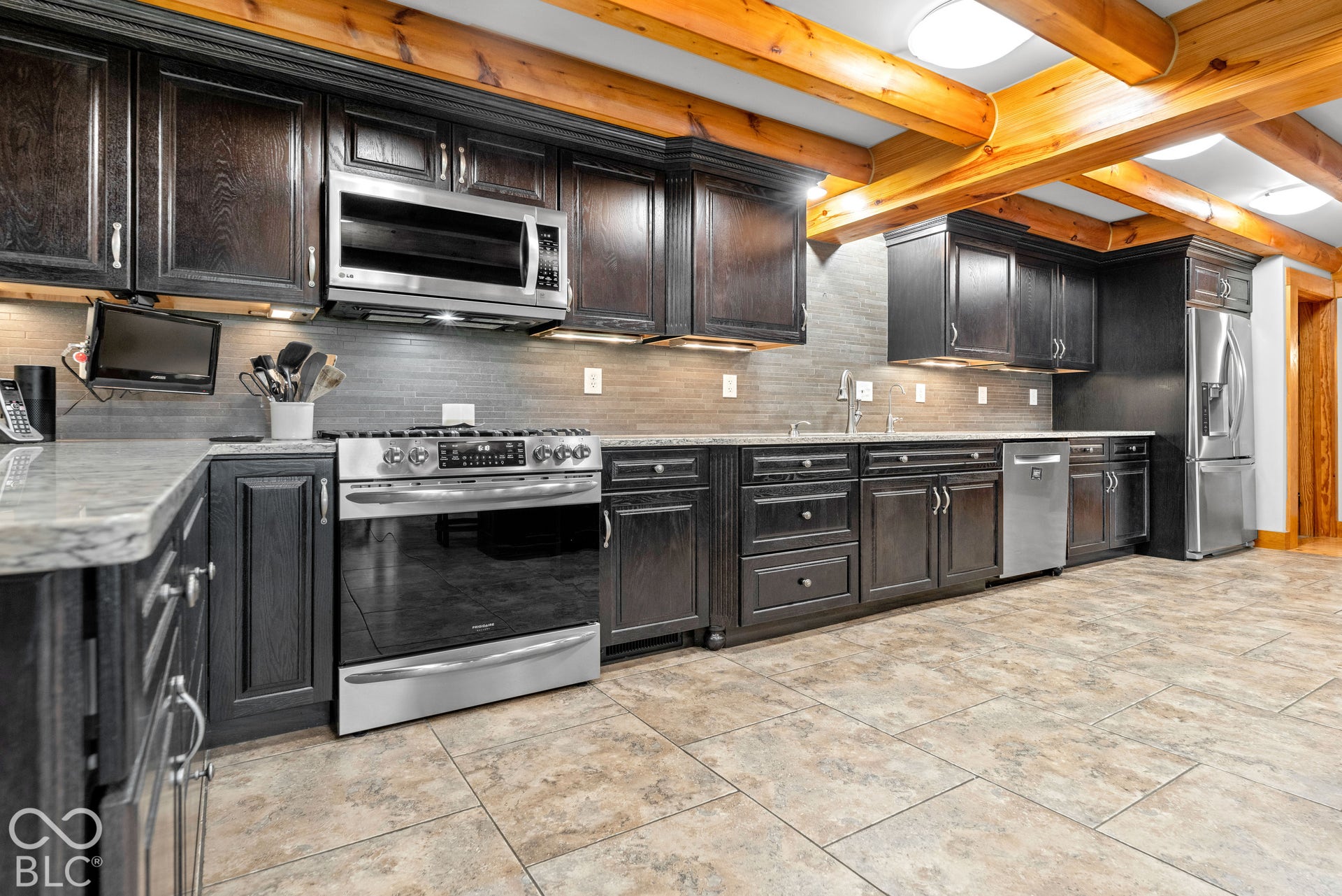 Dark wood cabinetry, stainless steel appliances, stone countertops, tiled backsplash, and exposed wood ceiling beams