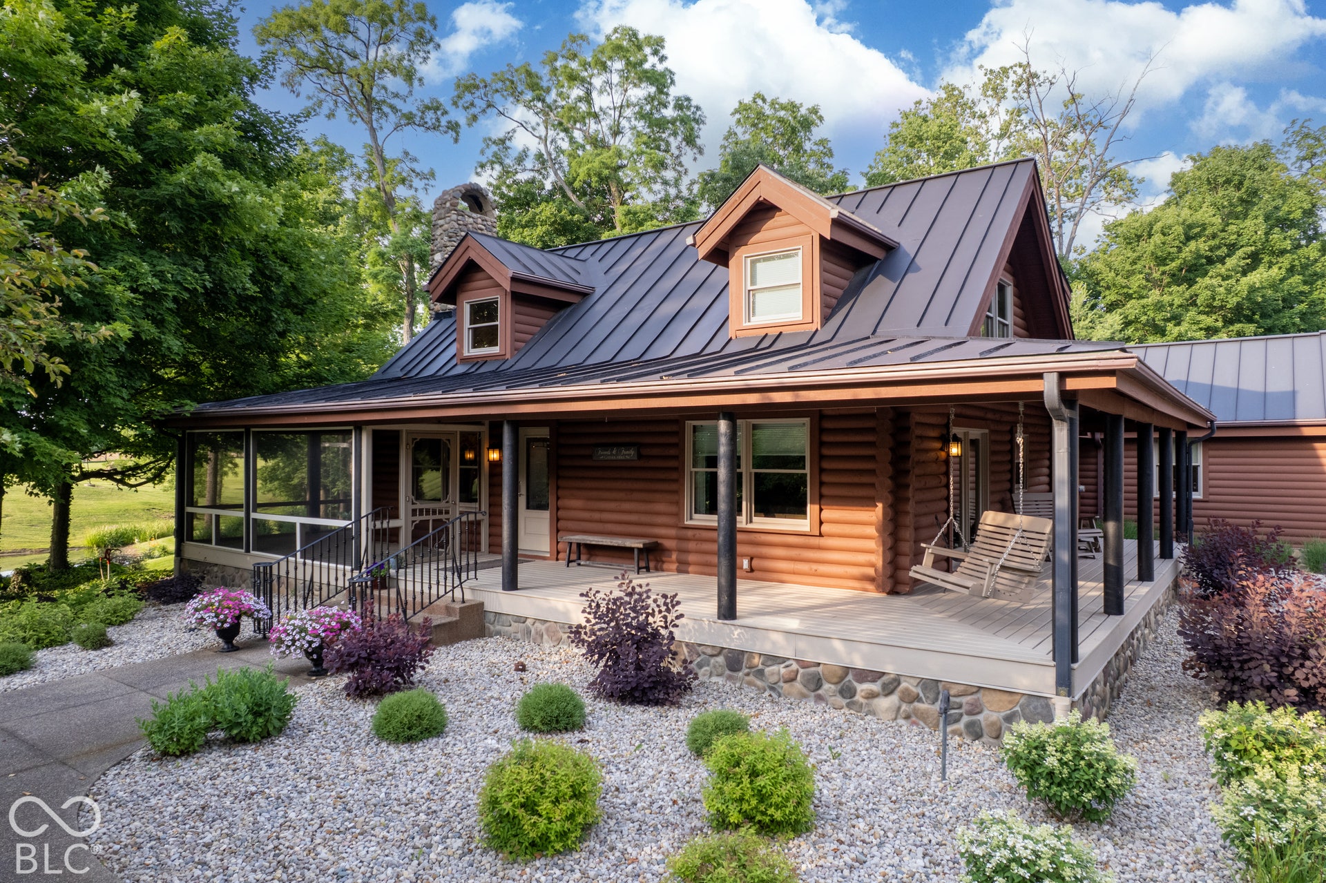 Log cabin exterior with a metal roof, dormer window, full-length covered porch, screened porch, and stone-clad foundation