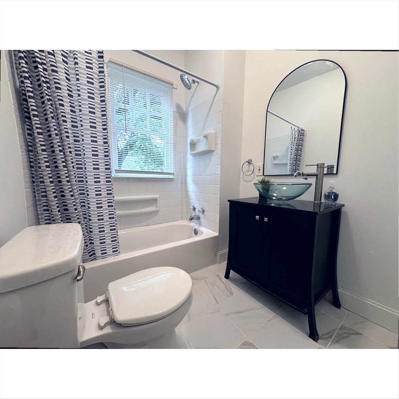 Bathtub with white tile surround, window with blinds, dark wood vanity with vessel sink, and arched mirror